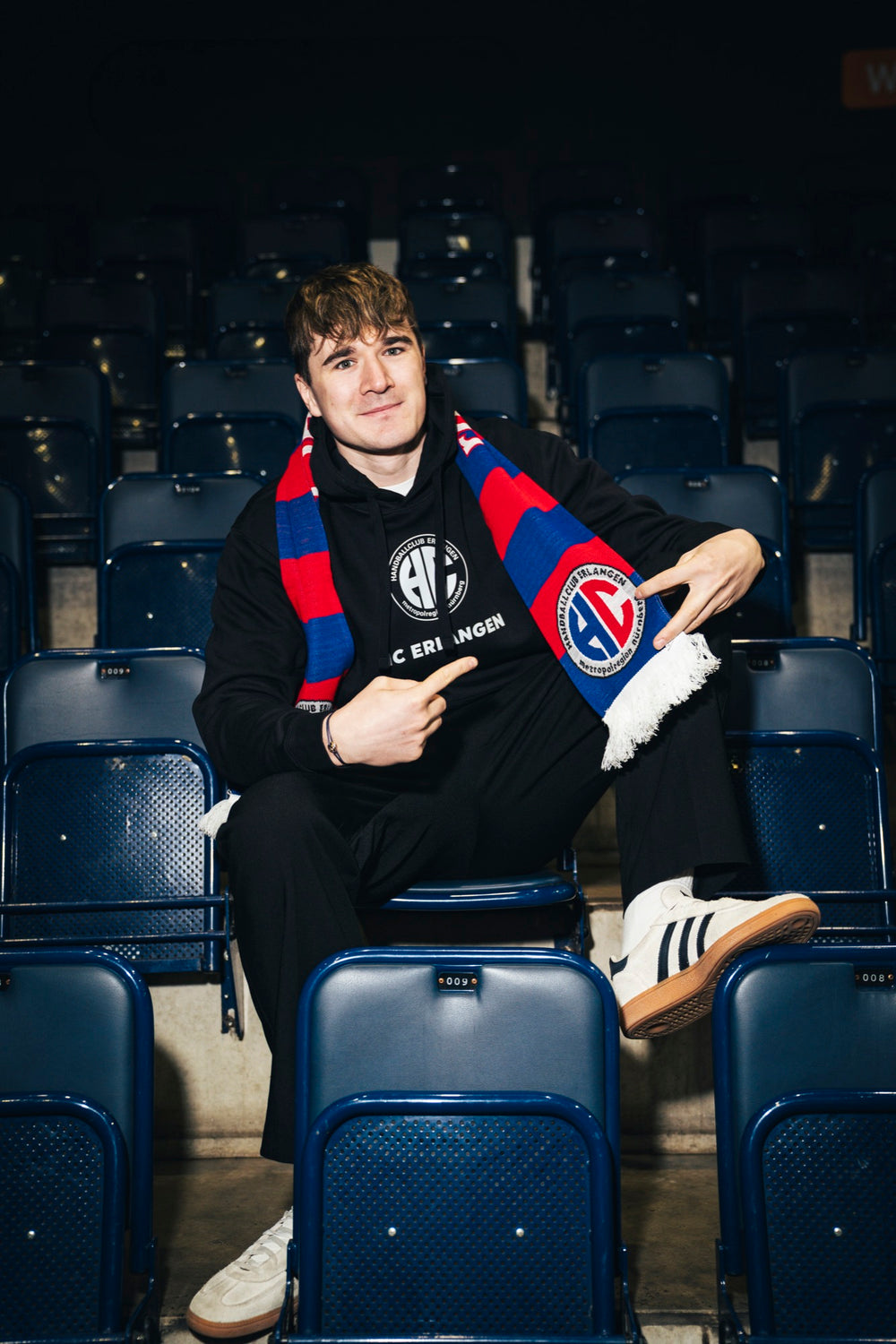 Yannik Bialowas im schwarzen HC Erlangen Classics Pullover mit dem Schal rot-blau auf der Tribüne
